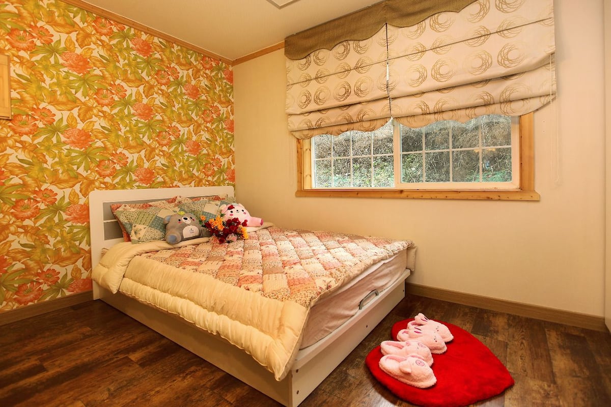 Une chambre avec un papier peint à motifs floraux, un lit recouvert d’une courtepointe, une fenêtre avec des rideaux et un tapis rouge.