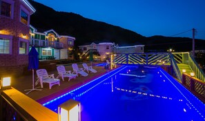 Zona piscina con lettini e un pontile in legno, circondata da edifici illuminati.