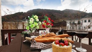 Una preparación para picnic con pan, pastel, fresas, copas de vino y flores sobre una mesa de madera, con vistas a la montaña.