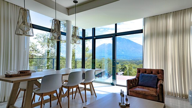 Uma área de jantar moderna com uma mesa grande, cadeiras e vista para uma montanha através de amplas janelas.