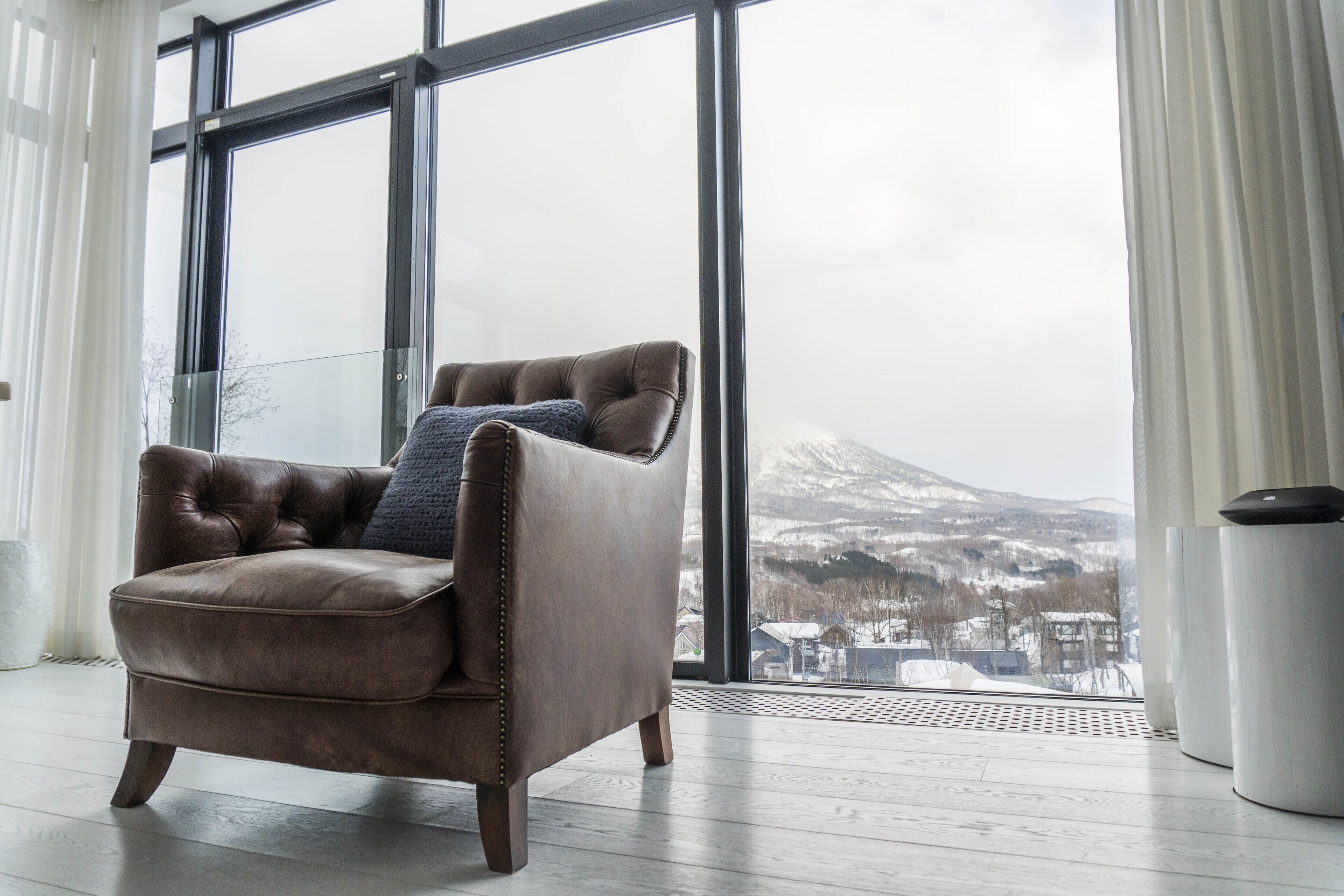 Un sillón de cuero marrón con un cojín azul en una habitación con ventanas amplias que dan a una montaña nevada.