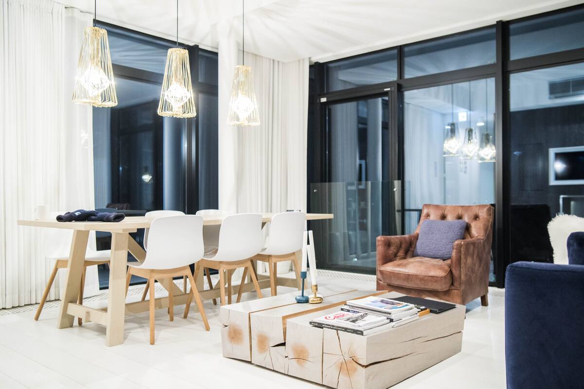 A modern living room with a dining area, featuring a wooden table, white chairs, a leather armchair, and a coffee table with magazines.