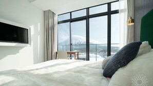 Quarto com uma janela grande, cama com roupa de cama branca e azul, televisão na parede e uma pequena mesa com uma cadeira.