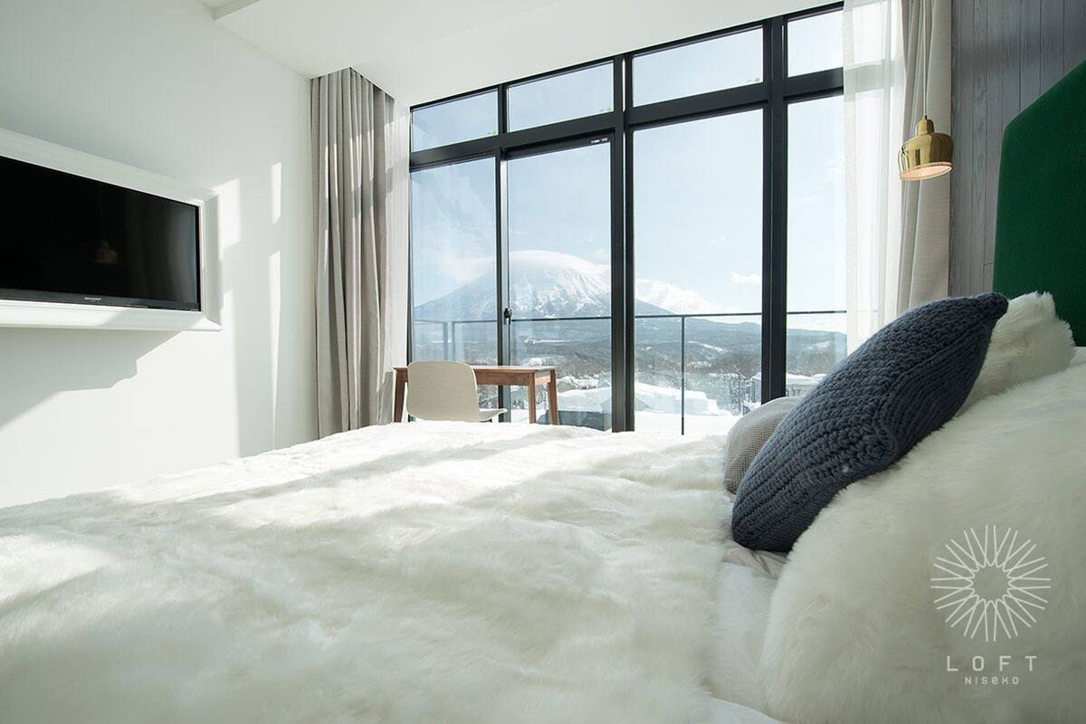 A bedroom with a large window, a bed with white and blue bedding, a TV mounted on the wall, and a small table with a chair.