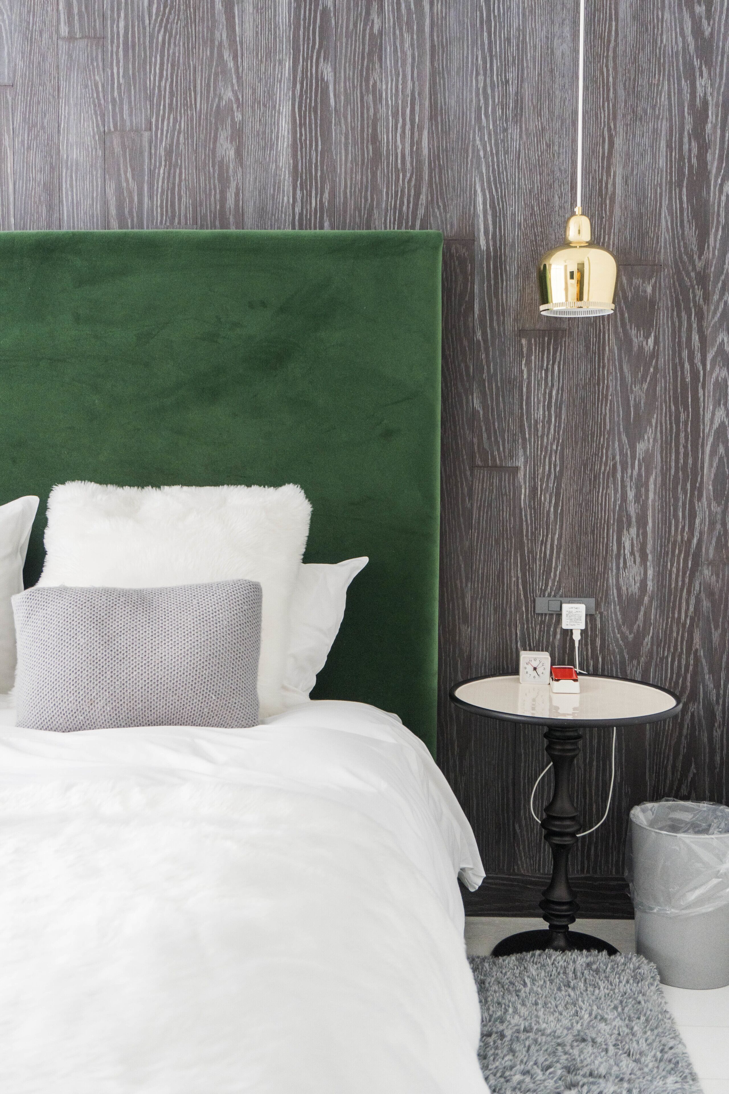 Un dormitorio ordenado con cabecera verde, ropa de cama blanca y una mesita de noche con lámpara.