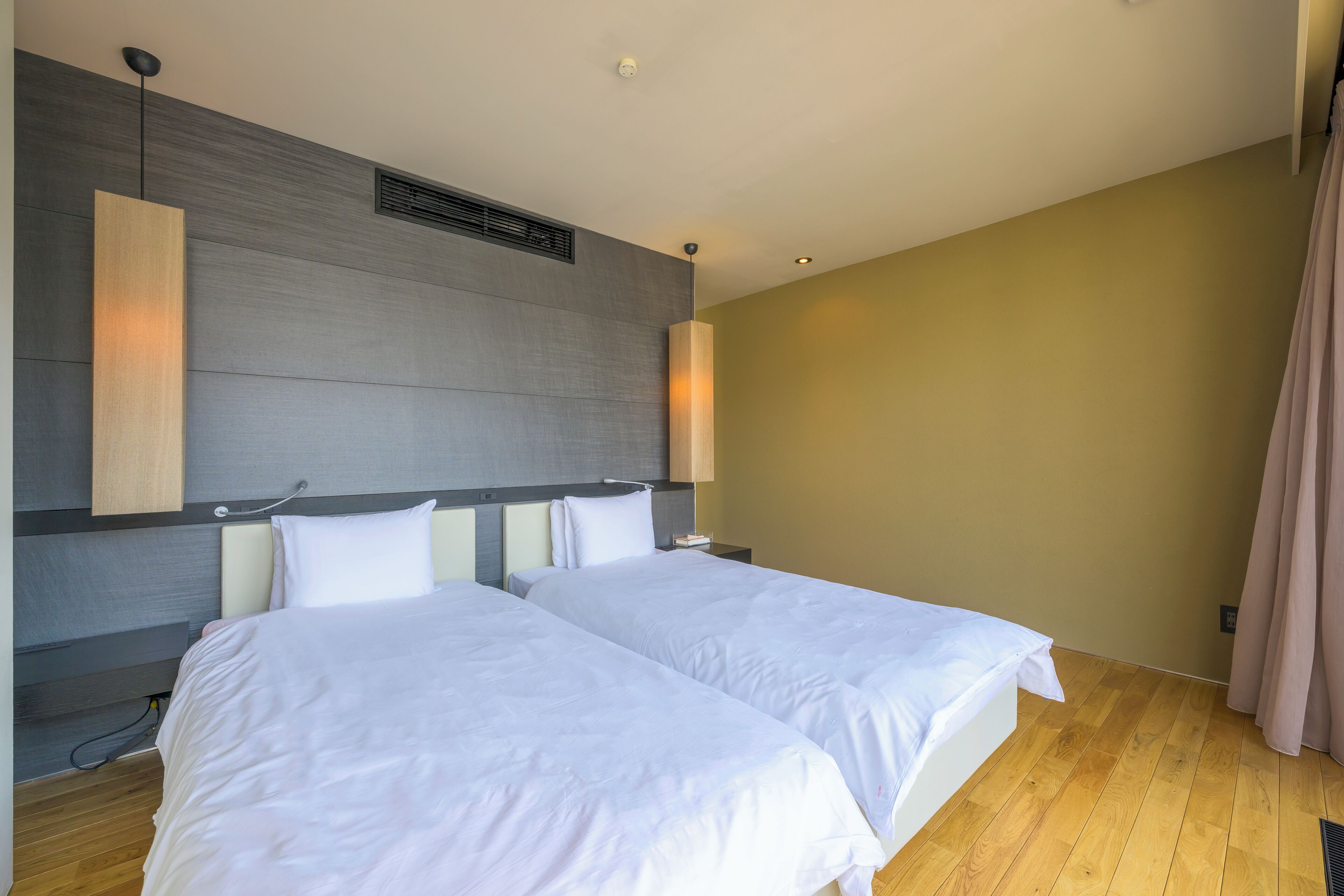 A hotel room with two beds, a wooden floor, a grey accent wall, and a beige wall.