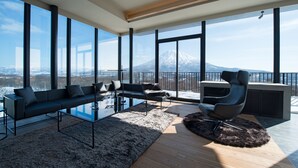 A modern living room with a glass coffee table, a sofa, and a chair, large windows offering a mountain view. - MUSE Niseko (Kutchan)