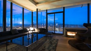 A modern living room with a glass coffee table, a sofa, and a fireplace. Large windows offer a view of the cityscape and a mountain. - MUSE Niseko (Kutchan)