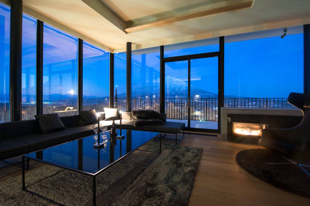Una sala moderna con una mesa de centro de vidrio, un sofá y una chimenea. Ventanas amplias ofrecen vistas a la ciudad y a la montaña.