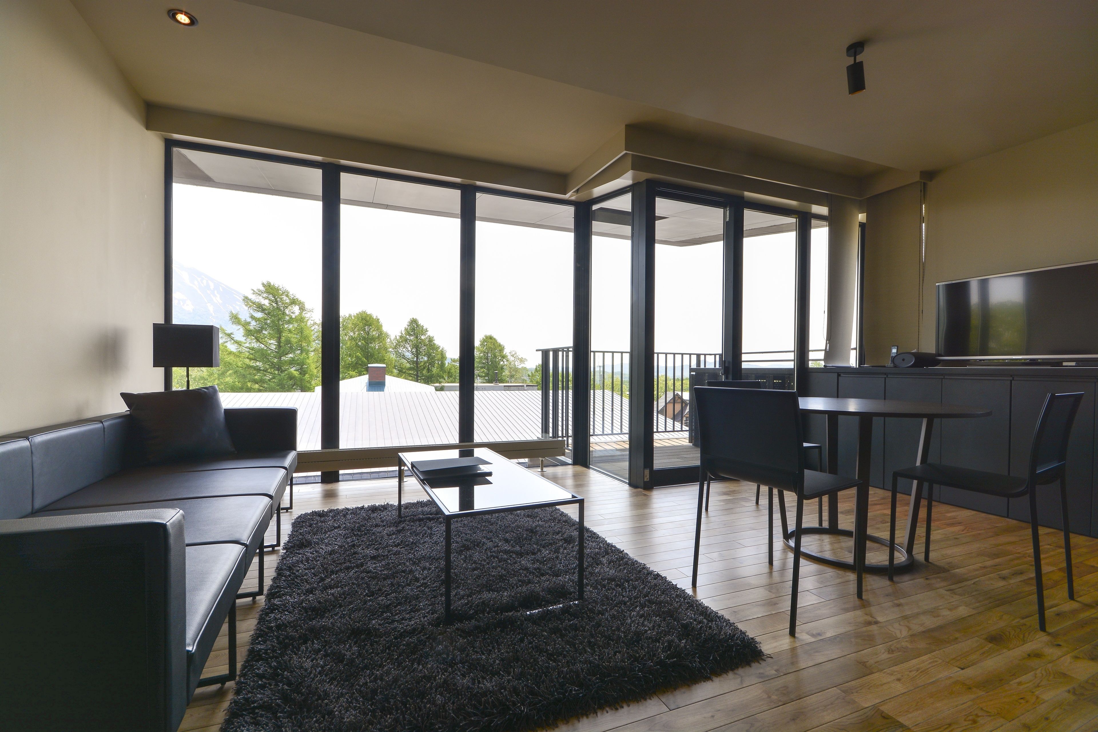 A modern living room with a sofa, a coffee table, and a TV. There is a balcony with a view of the mountains.