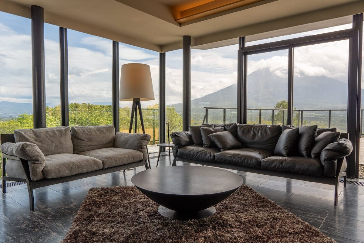 Una sala de estar moderna con dos sofás, una mesa de centro y una lámpara de pie, con un gran ventanal que ofrece vistas a la montaña.