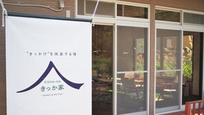 A building with a large banner displaying Japanese text, a wooden deck, and a view of a garden through the windows. - Kikkake Green and Mt.Fuji - Hostel (Fujiyoshida)