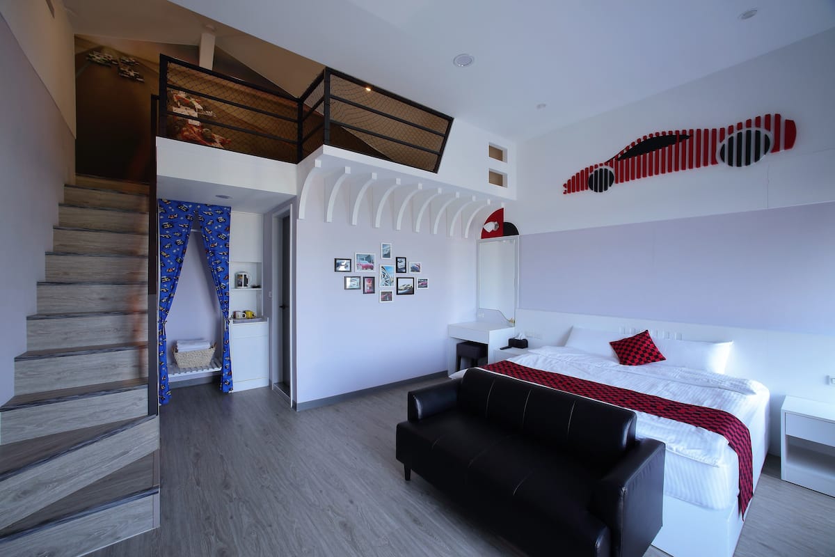 A modern hotel room with a large bed, a black leather sofa, a desk, and a staircase leading to an upper level.