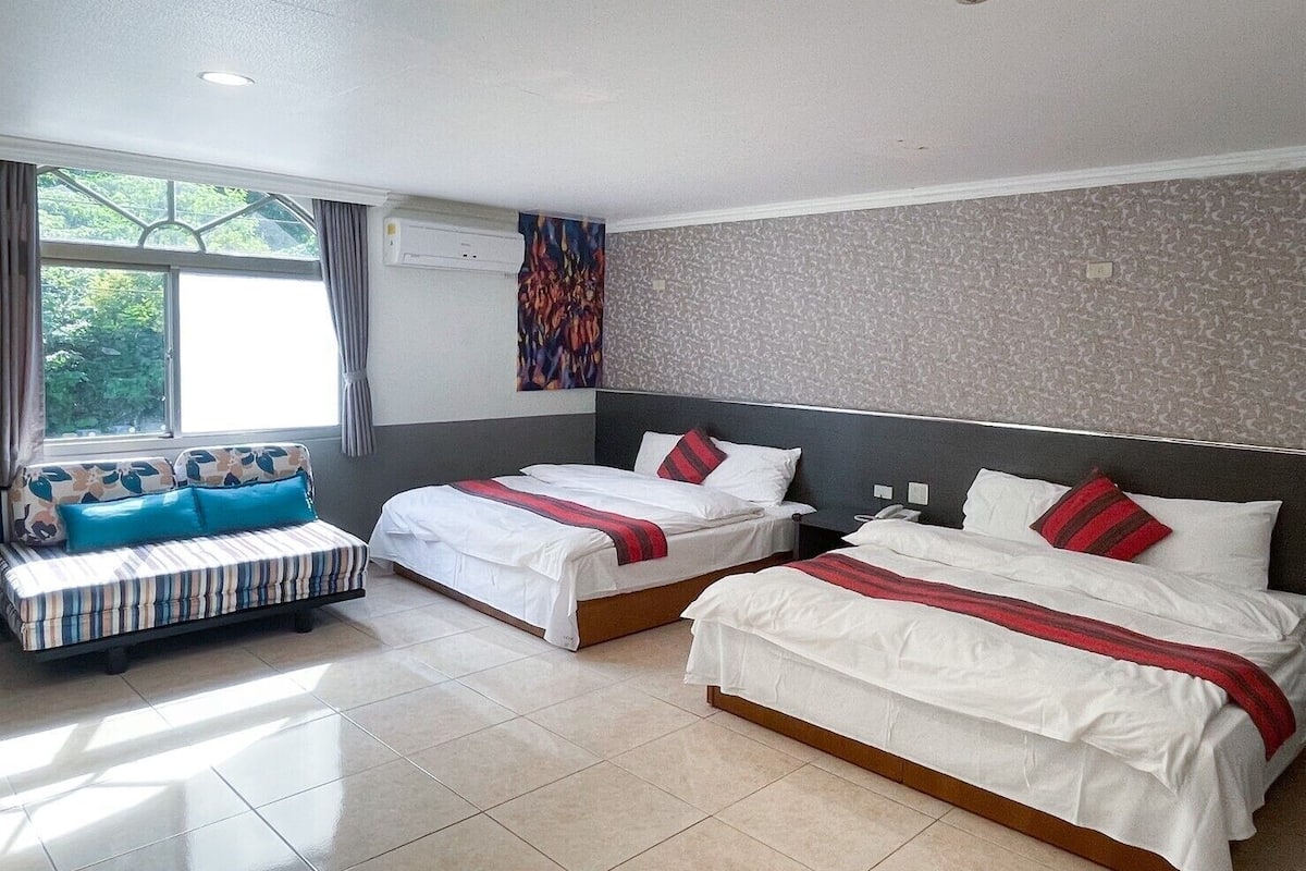 A hotel room with two beds, a sofa, a window with curtains, and a patterned wall.