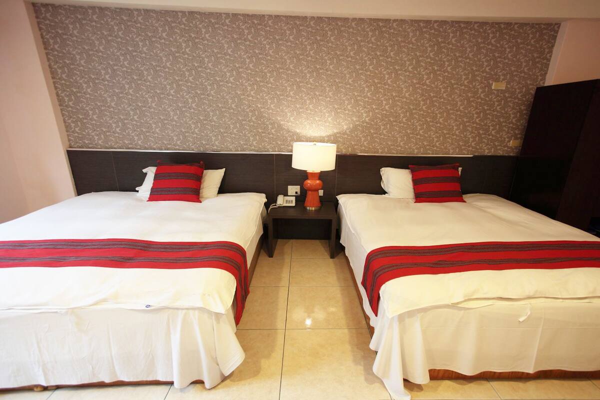 A hotel room with two beds, each with white bedding and red accents, a nightstand with a lamp, and a patterned wallpaper on the wall.