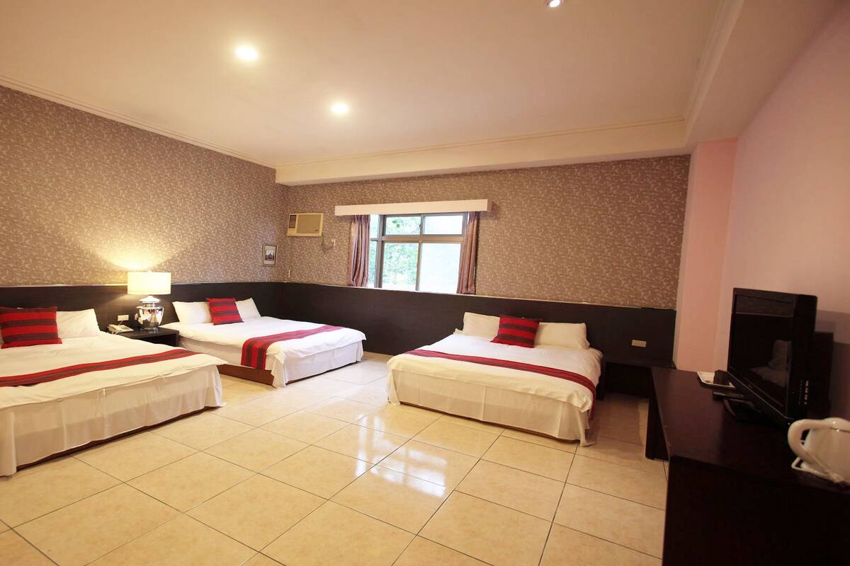 A hotel room with three beds, a TV, a desk, and a window.