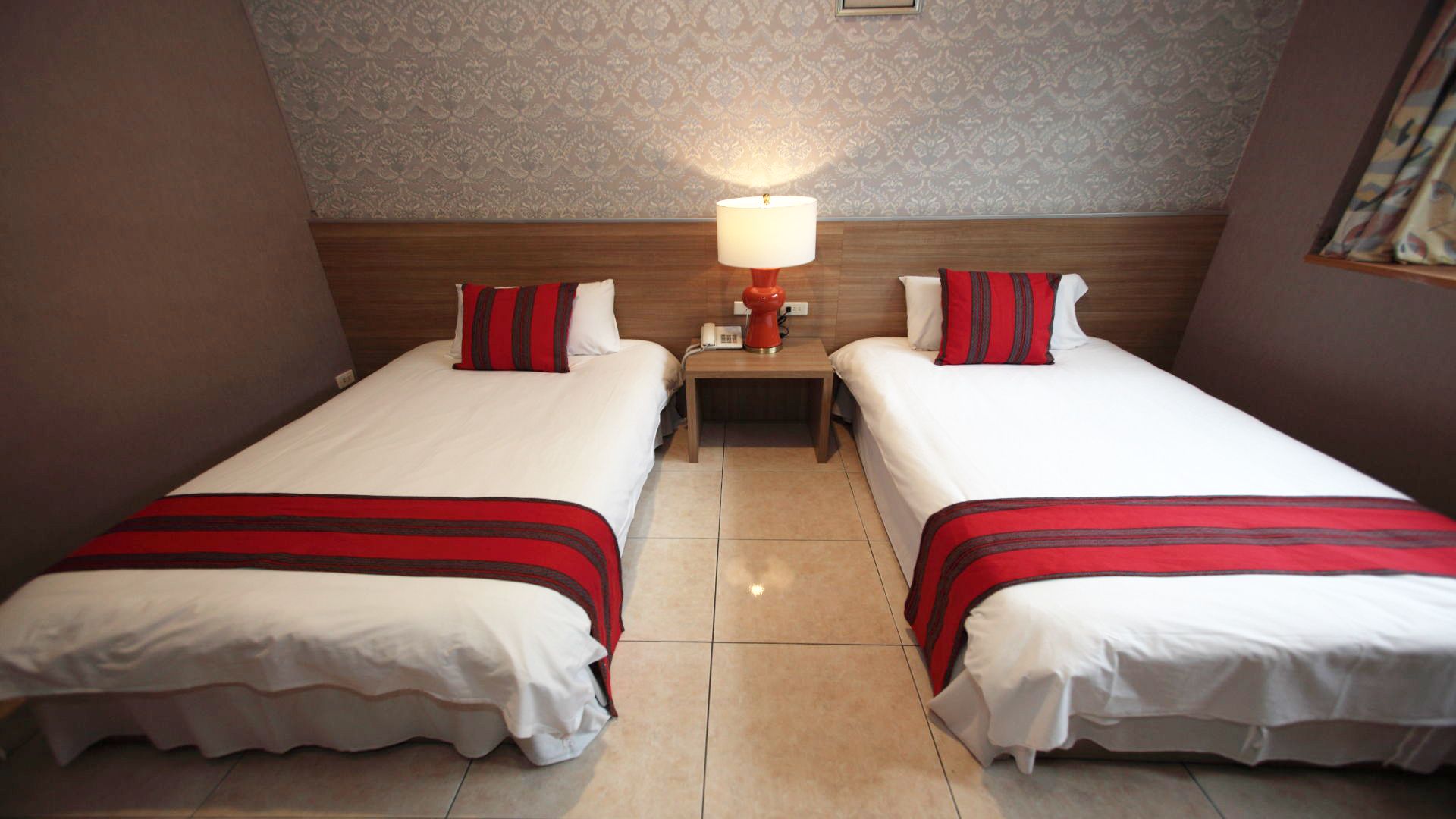 Una habitación de hotel con dos camas, cada una con una colcha a rayas rojas y blancas, una mesita de noche con una lámpara y un papel tapiz estampado en las paredes.