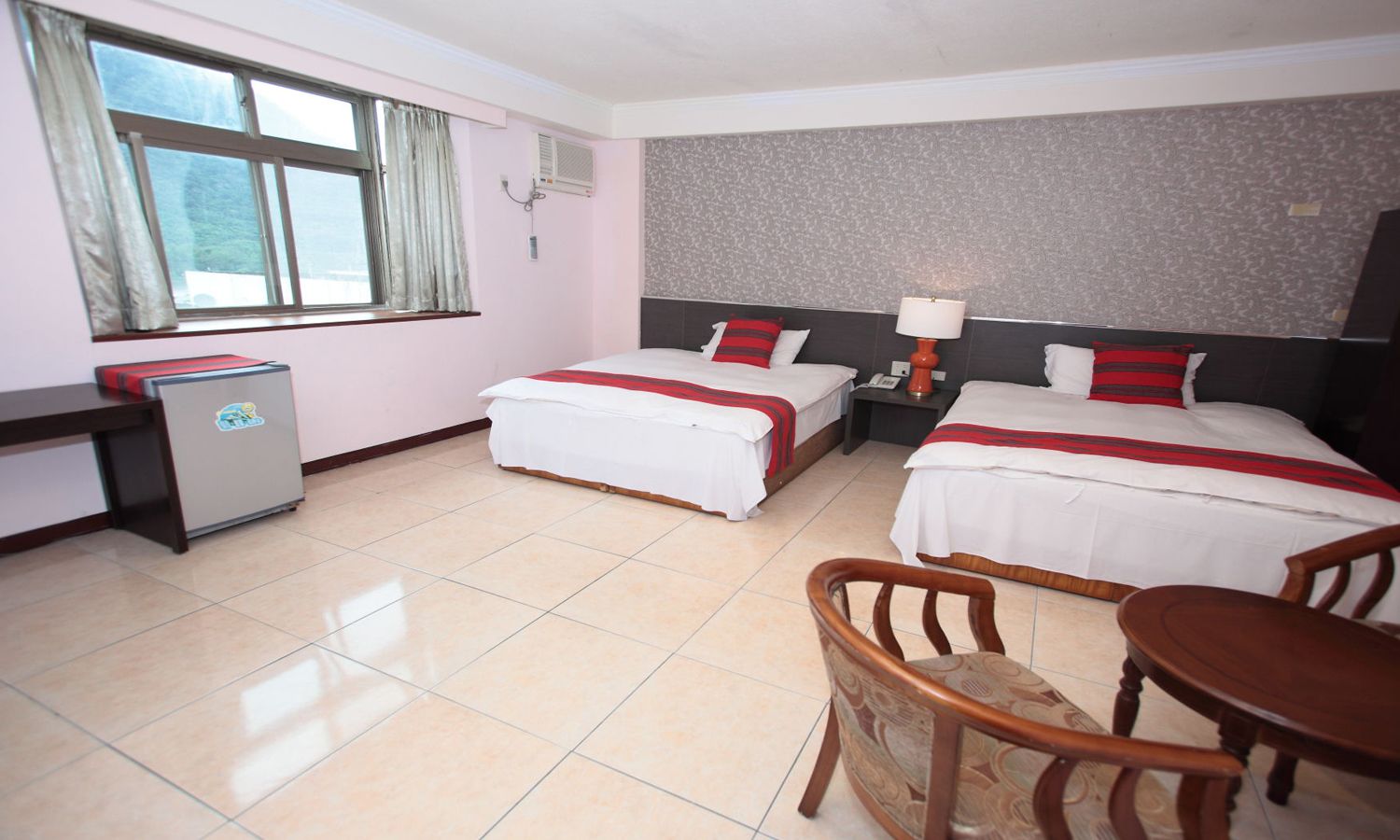 A hotel room with two beds, a desk, a chair, a table, and a refrigerator.