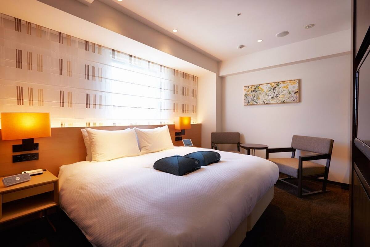 Quarto de hotel com uma cama grande, duas luminárias de cabeceira, uma mesinha, uma cadeira e um quadro na parede.