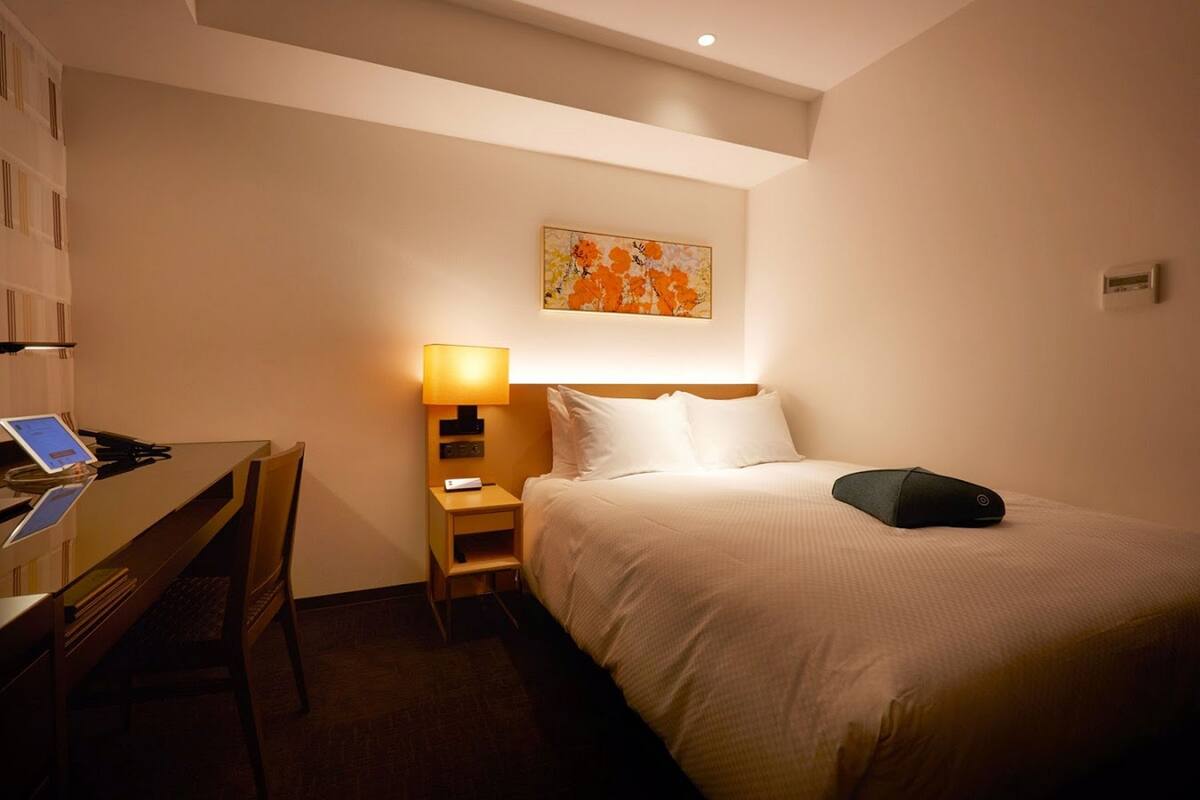 Quarto de hotel com uma cama, uma secretária com um laptop, uma mesa de cabeceira com abajur e um quadro na parede.