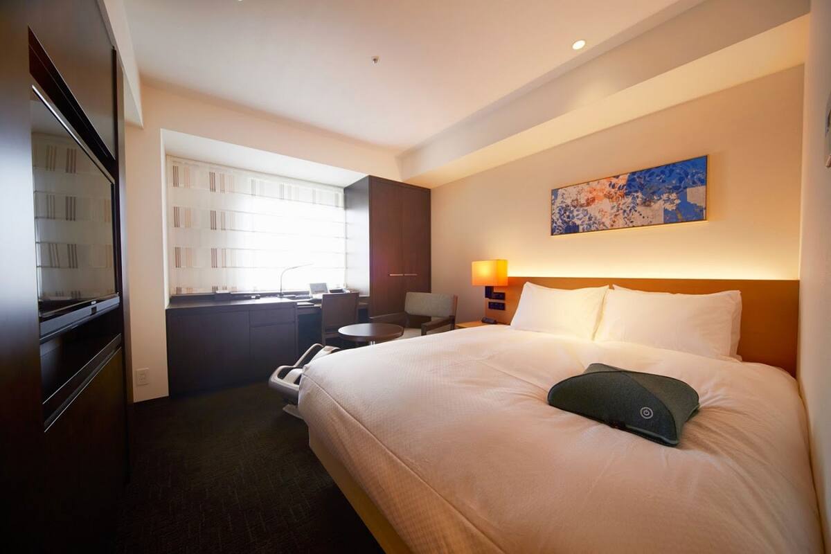 Quarto de hotel com uma cama grande, uma escrivaninha, uma cadeira, um abajur e um quadro na parede.