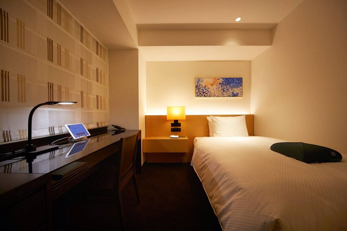 Quarto de hotel com uma cama, uma escrivaninha com um laptop, uma mesa de cabeceira com abajur e uma parede com um quadro.