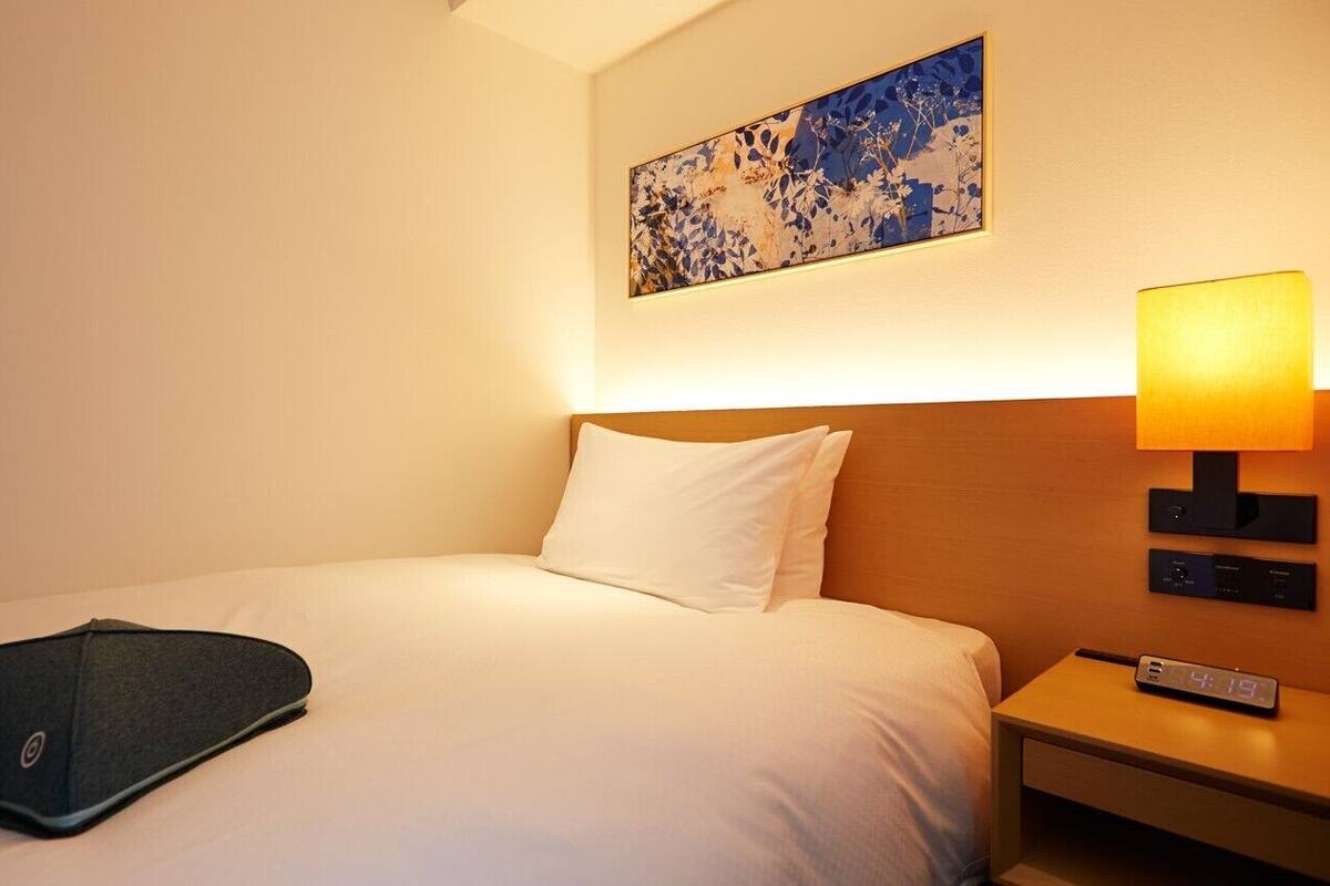 Quarto de hotel com cama, travesseiro, controle remoto, abajur e relógio.