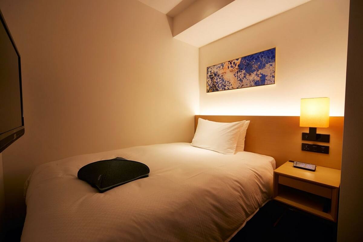 Quarto de hotel com cama, mesa de cabeceira com luminária, relógio digital e um quadro na parede.
