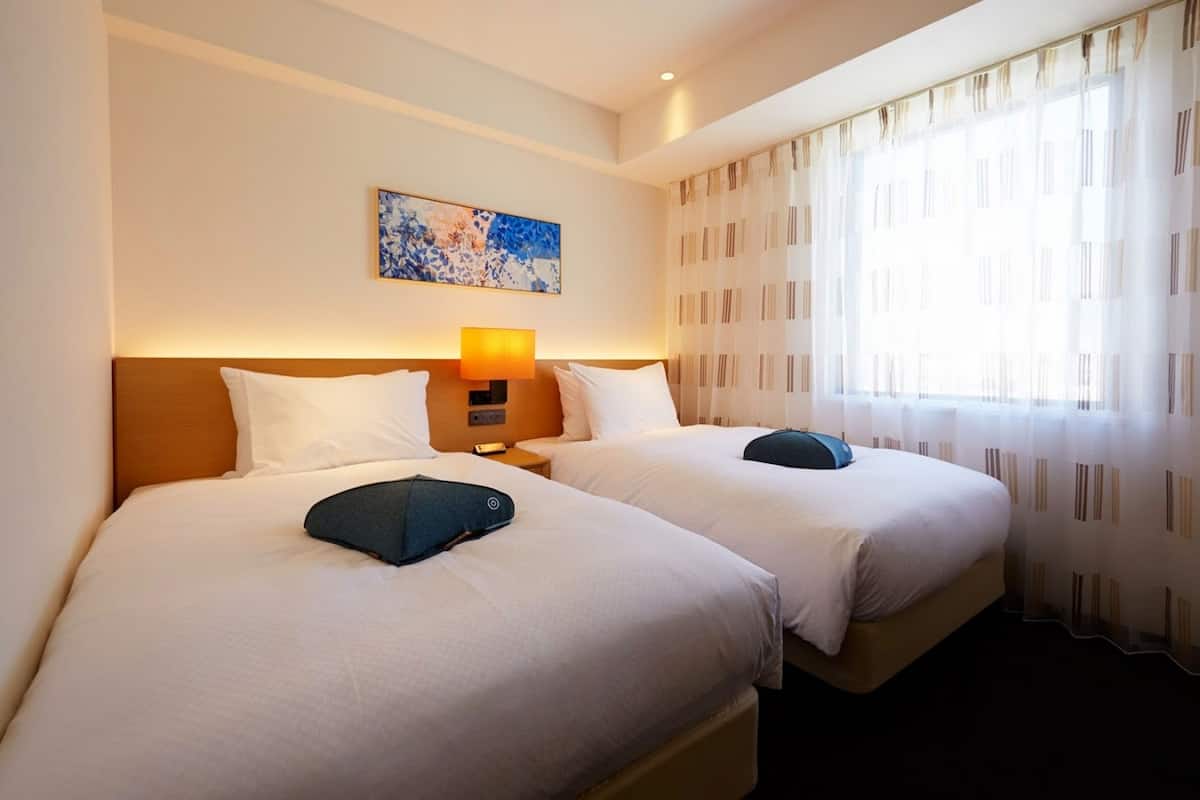 Quarto de hotel com duas camas, um quadro na parede, uma luminária e uma janela com cortinas.