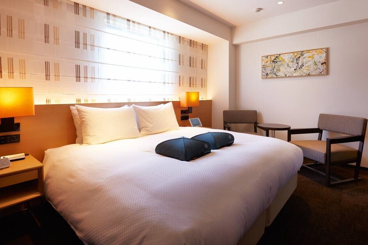 Quarto de hotel com cama grande, luminárias laterais, uma mesa pequena e uma cadeira.