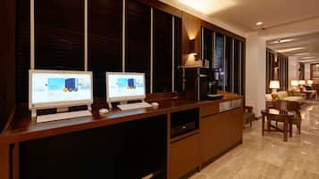 Un moderno hall de hotel con dos ordenadores de escritorio sobre un escritorio de madera, una máquina de café y una zona de estar con sofá y sillones.