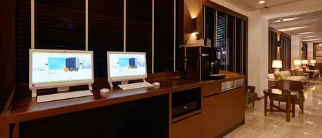 Un moderno hall de hotel con dos ordenadores de escritorio sobre un escritorio de madera, una máquina de café y una zona de estar con sofá y sillones.