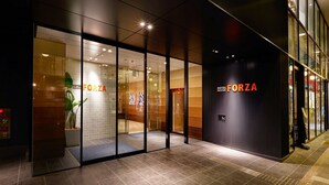 Une entrée d'hôtel moderne avec des portes en verre, un comptoir de réception bien visible et le nom « HÔTEL FORZA » affiché sur la façade.