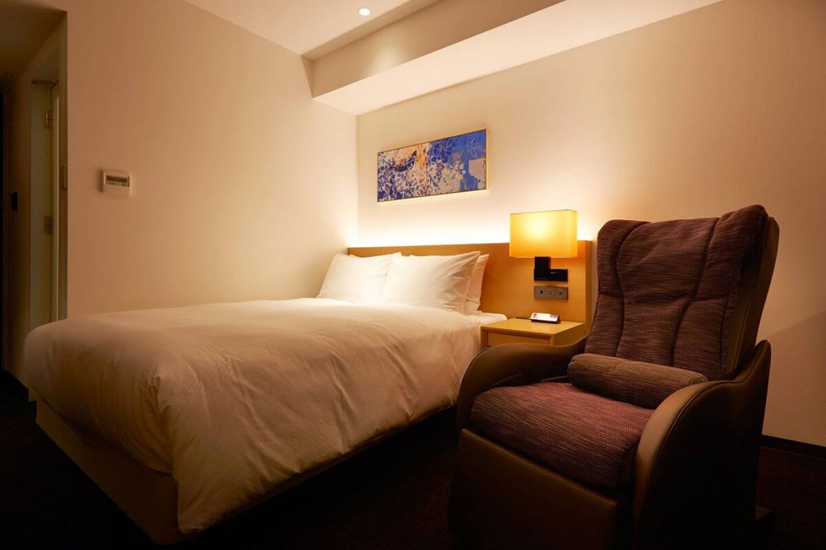 Quarto de hotel com cama, uma cadeira, mesa de cabeceira com luminária e um quadro na parede.