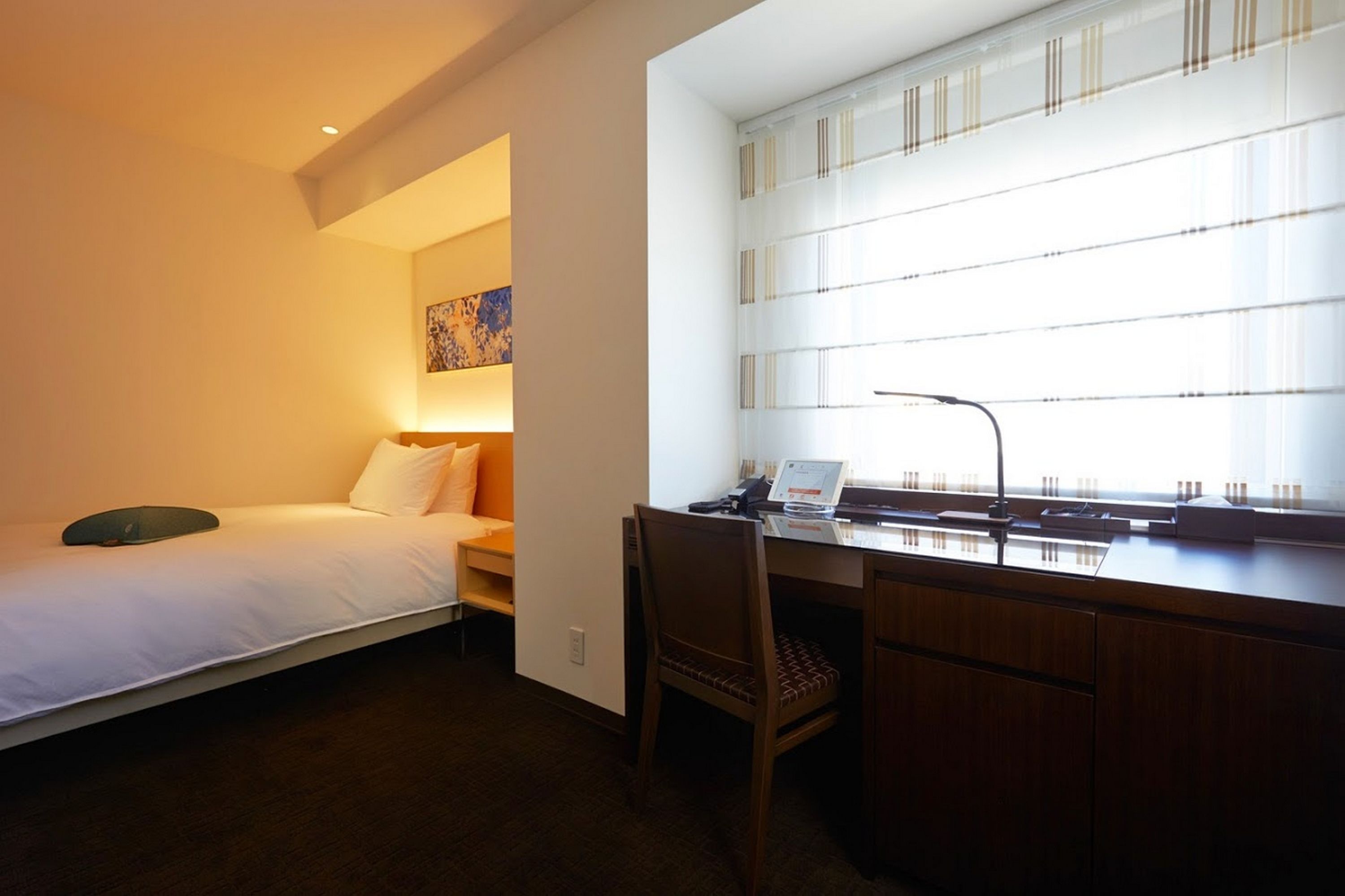 A hotel room with a bed, a desk with a laptop, and a window with blinds.