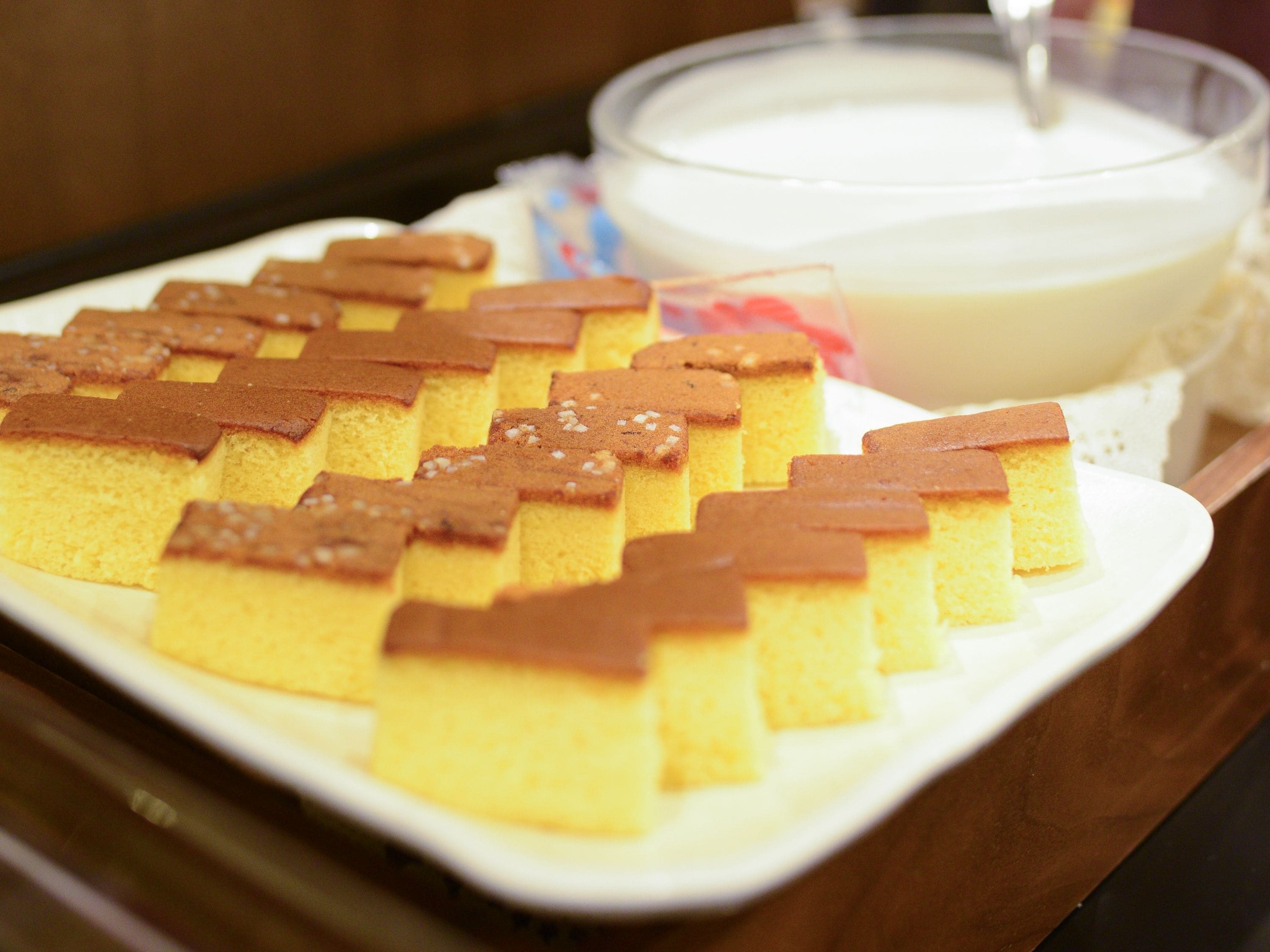 Un piatto di torta paradiso a cubetti con una copertura caramellata, accompagnato da una ciotola di crema bianca.