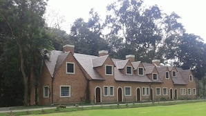 Uma fileira de casas de tijolo com telhados inclinados e várias chaminés, cercada por um gramado bem cuidado e árvores.