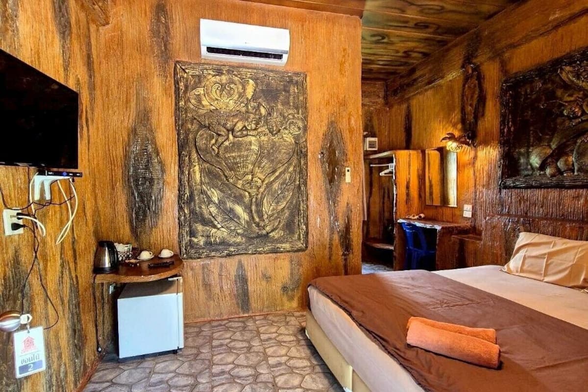 A room with a stone-tiled floor, a bed with a brown bedspread, a mounted TV, a small refrigerator, and a wall-mounted air conditioning unit.