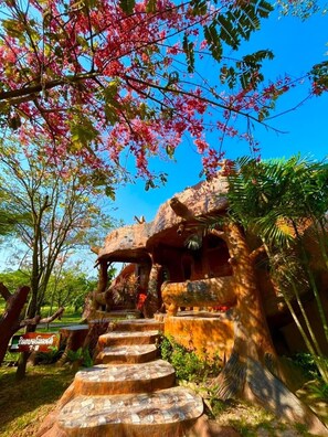 Una casa en un árbol con una escalera que lleva hasta ella, rodeada de vegetación exuberante y flores rosadas.