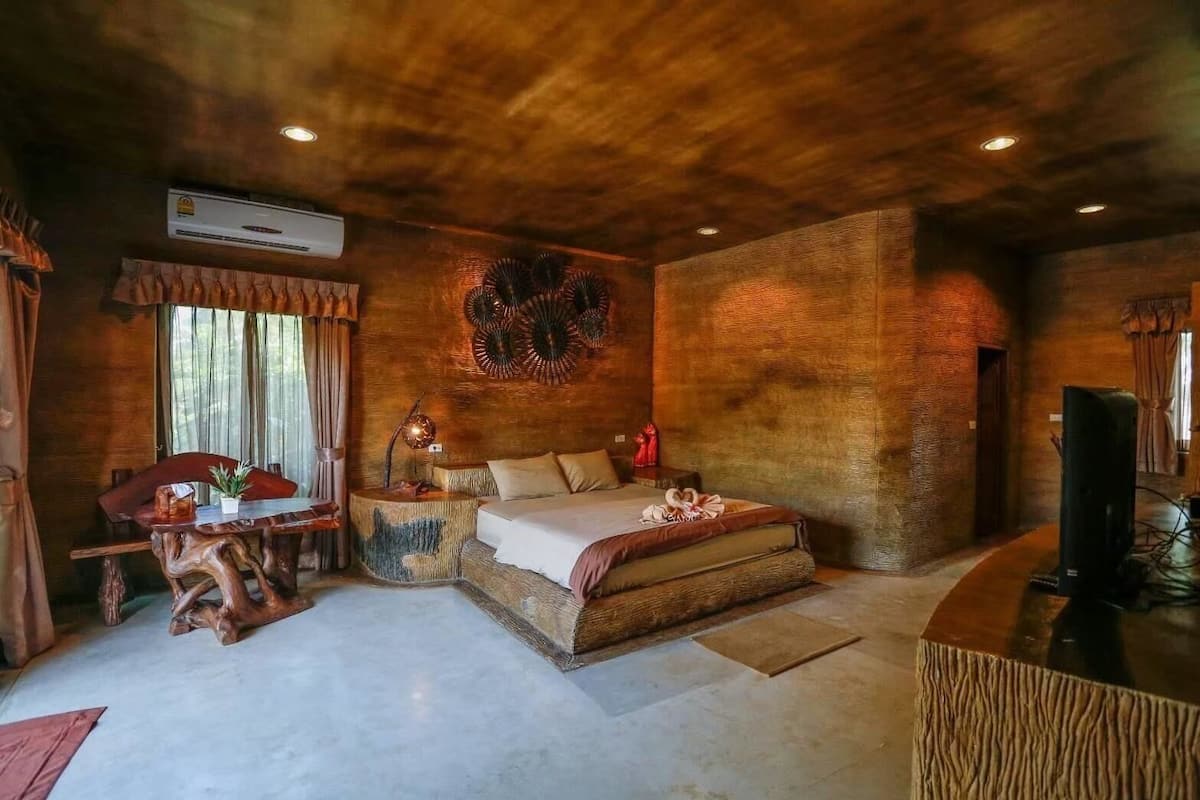 A rustic bedroom with a wooden ceiling, a bed with a wooden headboard, a dining table with chairs, and a television on a wooden stand.