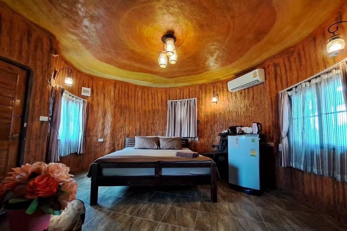 A wooden room with a bed, a small refrigerator, and a window with curtains.