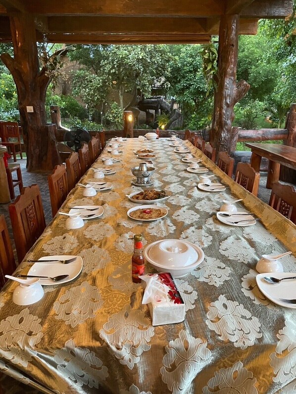 Una larga mesa de comedor dispuesta con platos, fuentes y cubiertos, rodeada de sillas de madera, con un entorno natural de árboles y una estructura de piedra.