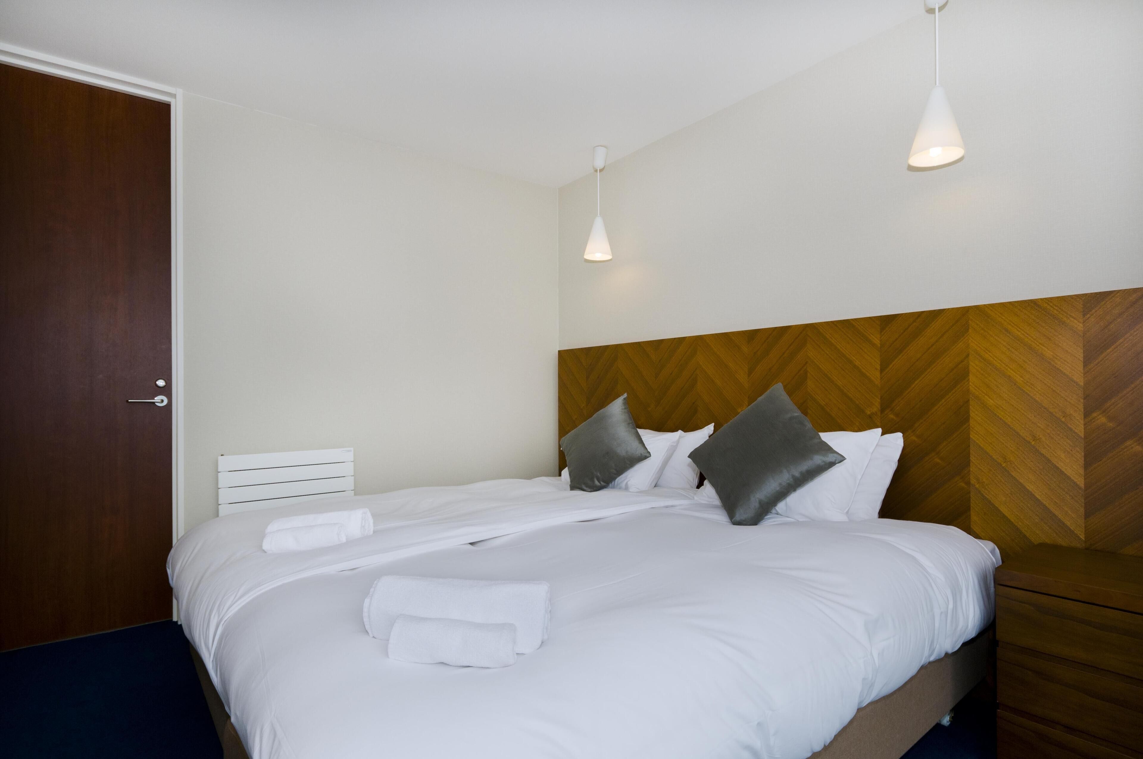 A neatly made bed with white linens and pillows, a wooden headboard, and two pendant lights.