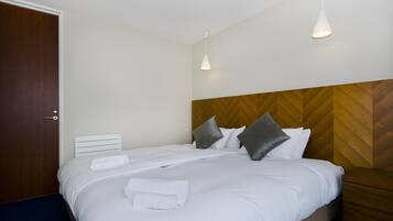 A neatly made bed with white linens and pillows, a wooden headboard, and two pendant lights.