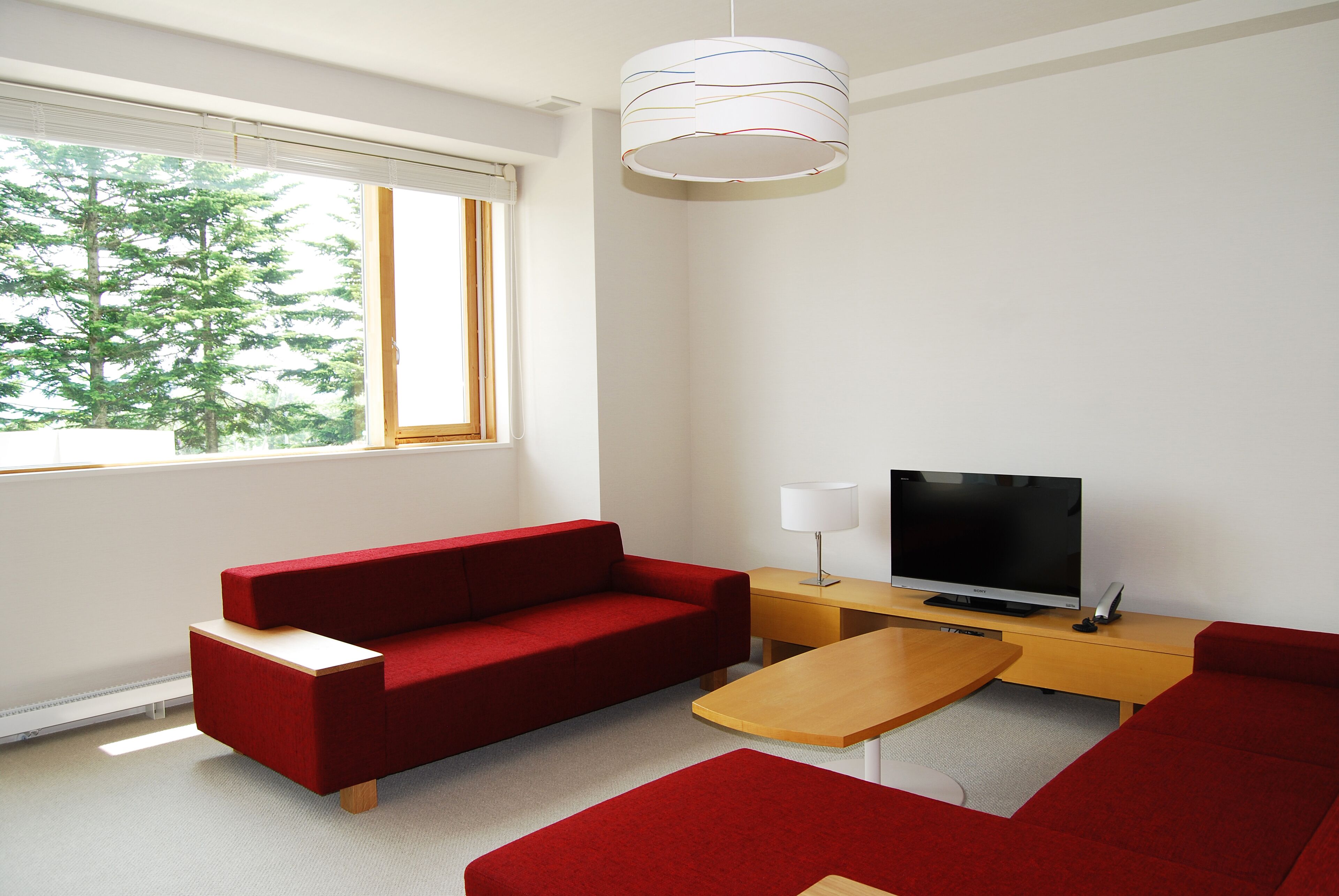 Ein modernes Wohnzimmer mit einem roten Sofa, einem Holztisch, einem Fernseher auf einem Holzschrank und einem großen Fenster mit Blick auf Bäume.