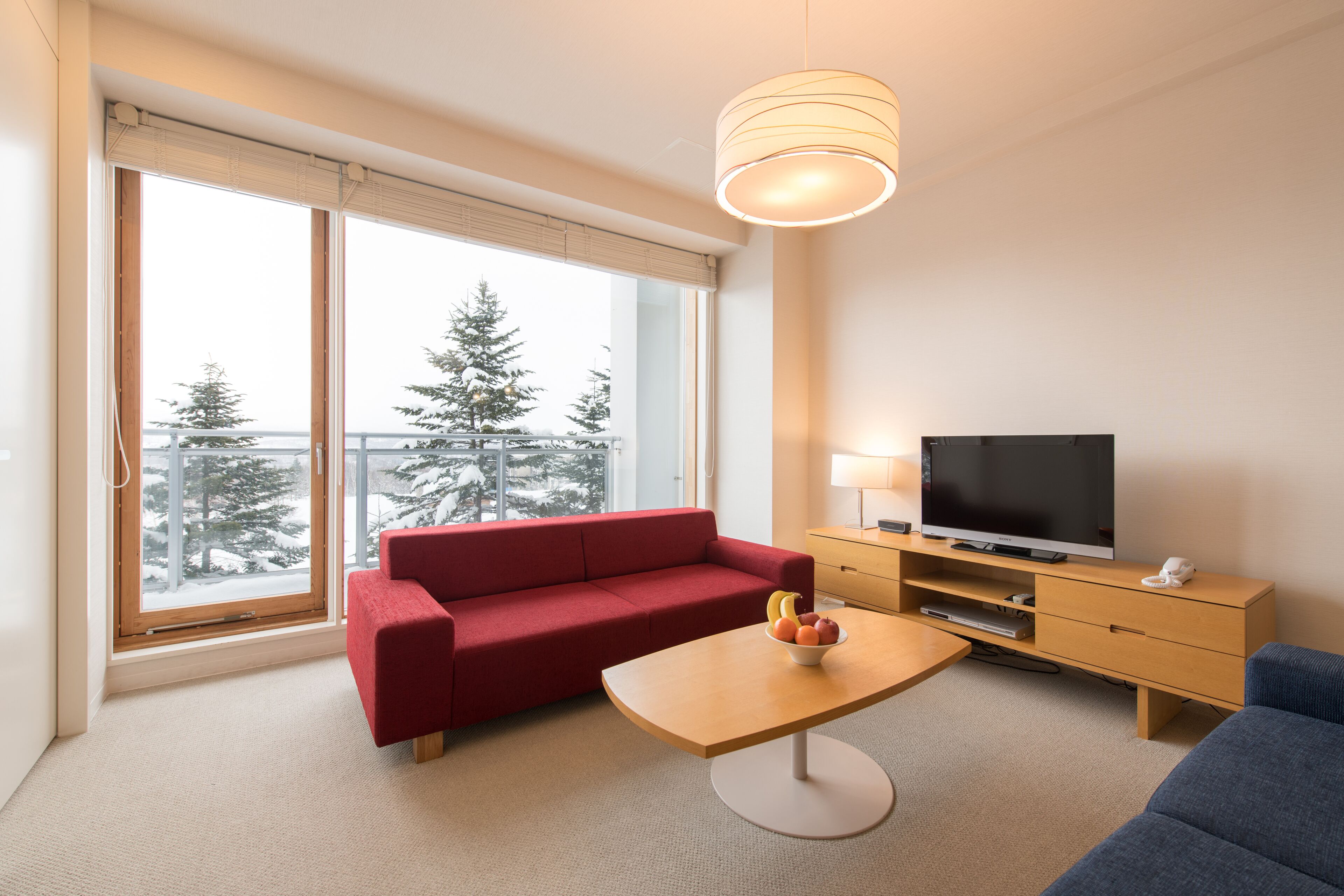 A modern hotel room with a red sofa, a wooden coffee table with fruit, a TV on a wooden stand, and a large window with a view of trees and a balcony.