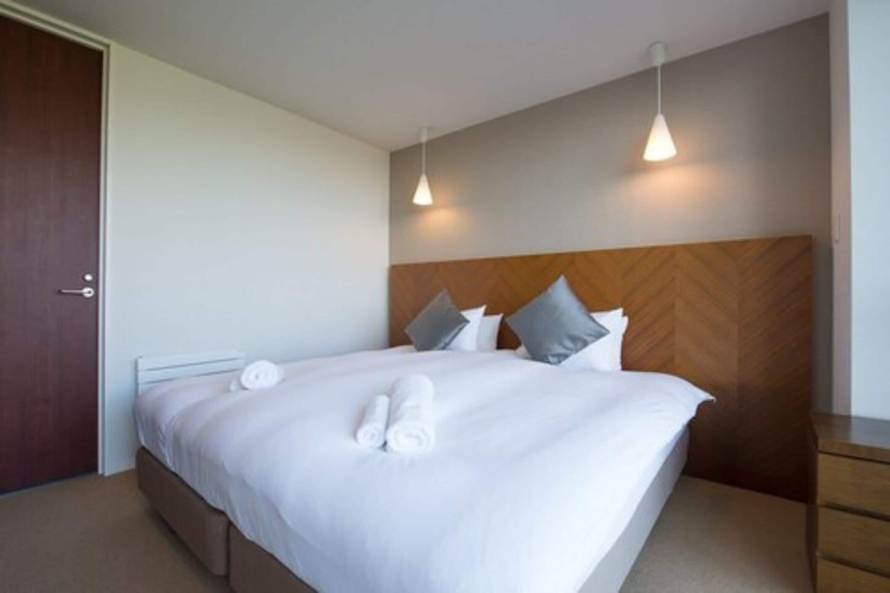 Una cama bien hecha con ropa de cama blanca y toallas enrolladas, un cabecero de madera y dos lámparas colgantes.