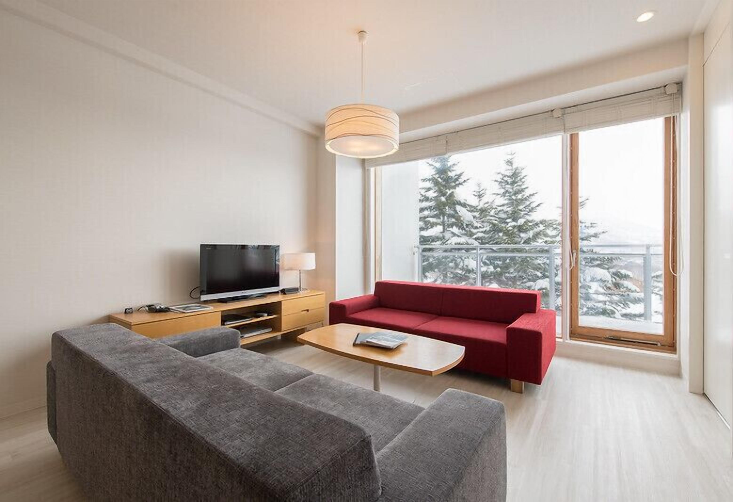 A modern living room with a grey sofa, a red couch, a television, and a large window with a view of snow-covered trees.