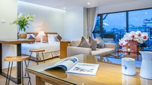 A modern hotel room with a bed, sofa, and a glass coffee table. There is a window with a city view, and a vase with flowers on the table. - Dream Hotel & Apartment (Hanoi)