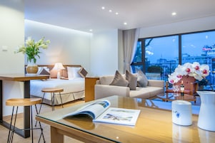 Quarto de hotel moderno com cama, sofá e mesa de centro de vidro. Há uma janela com vista para a cidade e um vaso com flores na mesa.